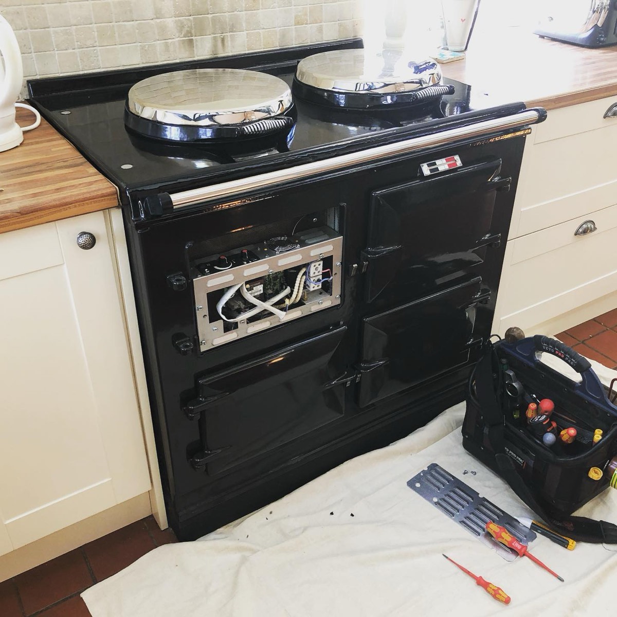 A black Aga with a front door off and its control box open, a tool bag and screwdrivers on a dust sheet beside it