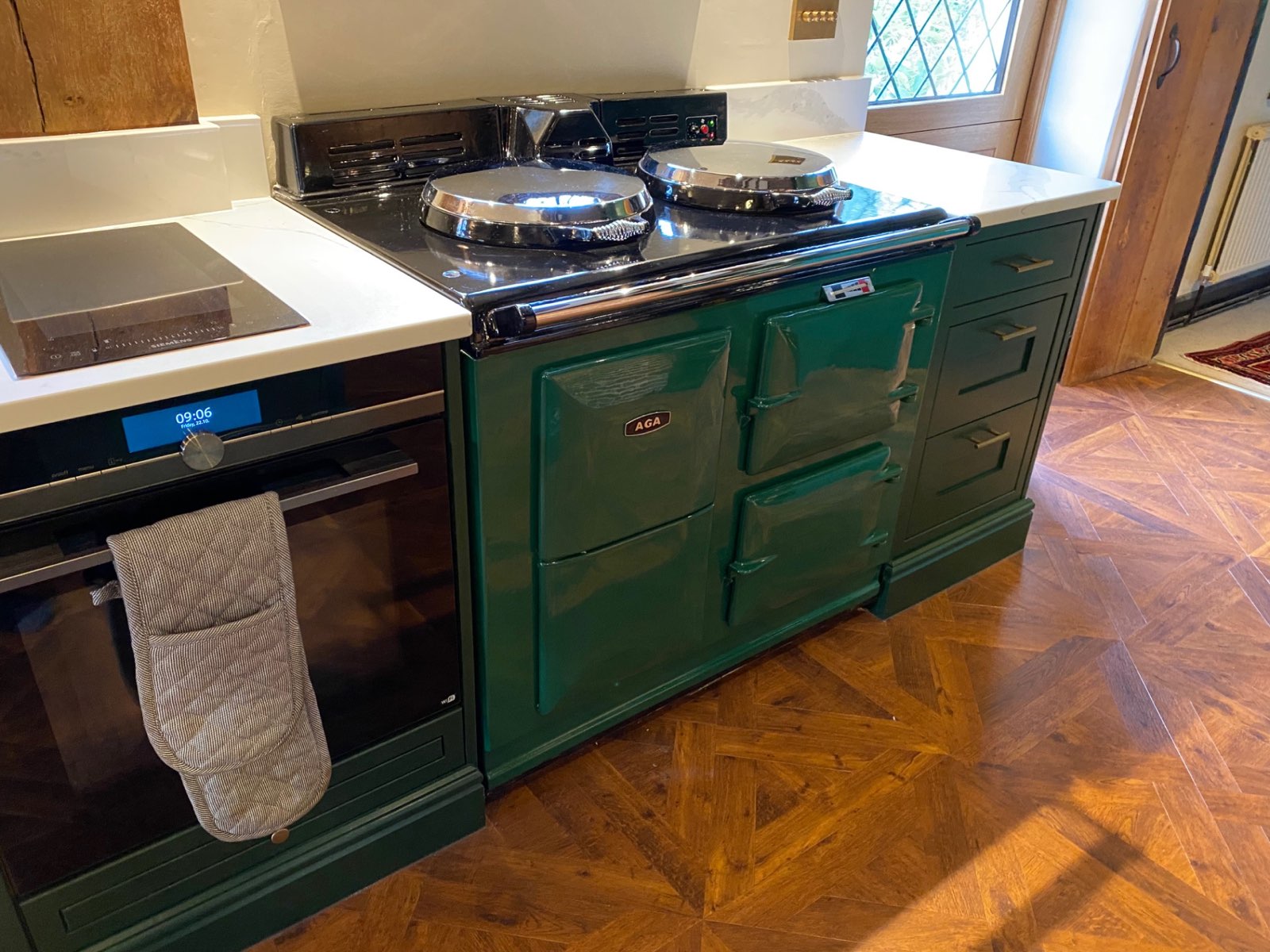 A refurbished racing-green Aga range cooker with polished chrome hotplate lids, fitted between green units on a parquet floor